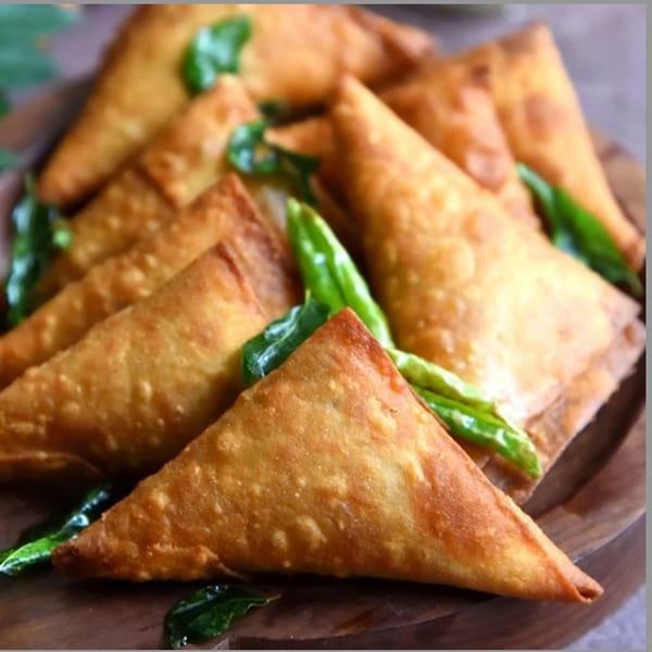 Golden-brown triangular samosas, garnished with green chilies and curry leaves, on a wooden platter.