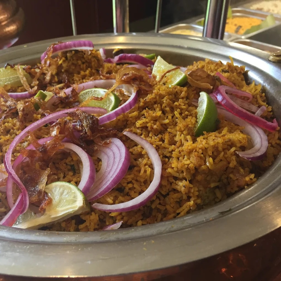 Close-up of Kaveri South Indian Restaurant's biryani, garnished with red onions, fried onions, and lime wedges.