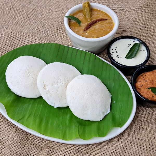 Three white idlis on a banana leaf with sambar, coconut chutney, and tomato chutney.