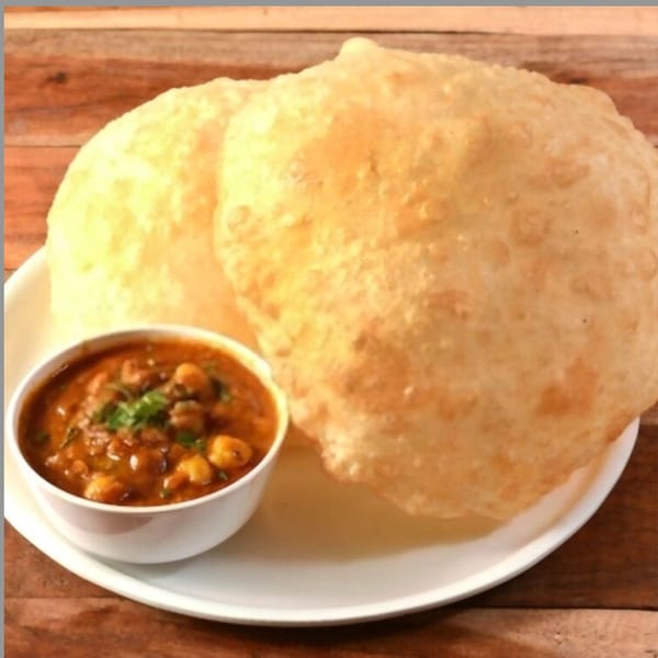 Two golden-brown poori breads on a white plate next to a bowl of chana masala.