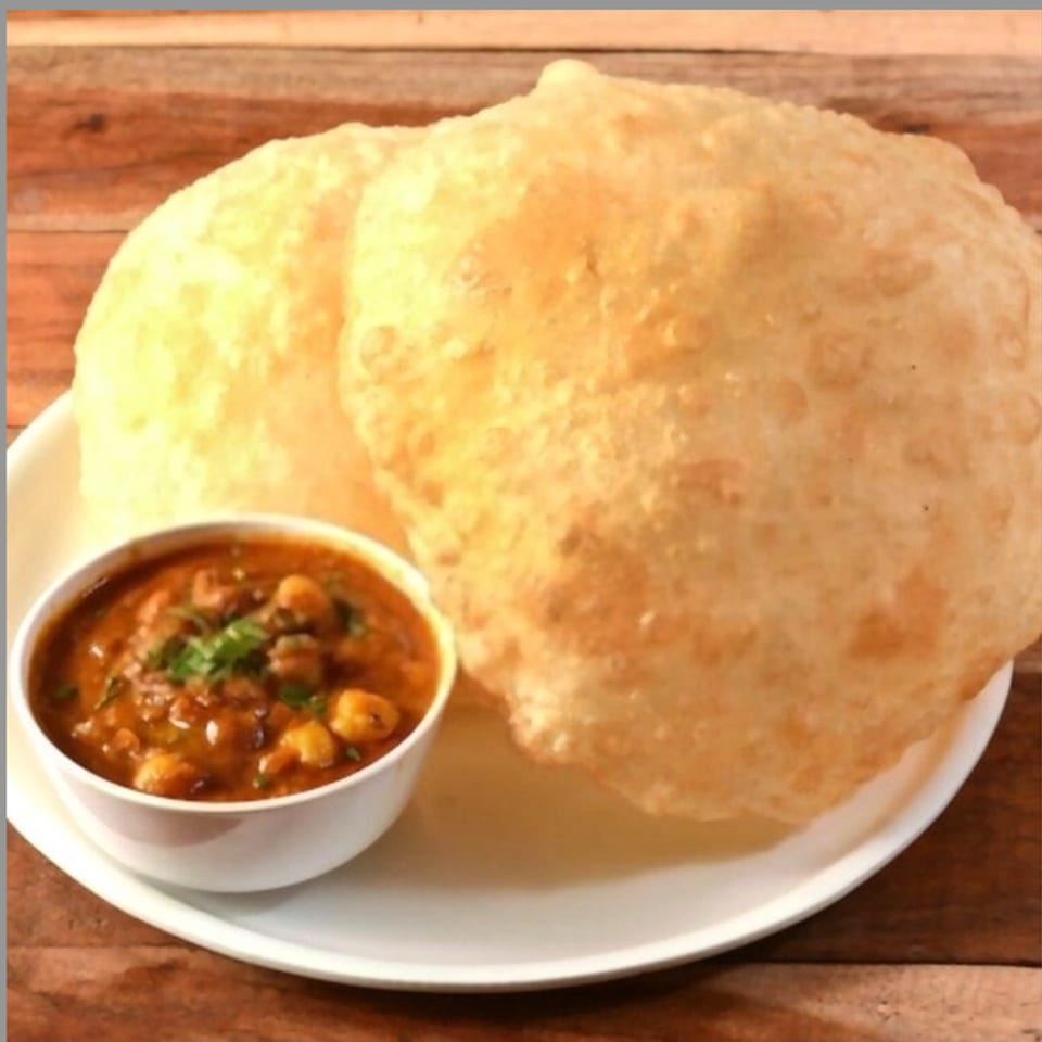 Two golden-brown poori breads on a white plate next to a bowl of chana masala.